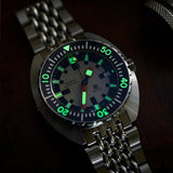 A close-up of a DOXA ARMY Automatic 42.50mm Watch 785.10.031.10 with a matching link bracelet, featuring a black dial with bright green and white luminous markers and hands, set against a dark background.