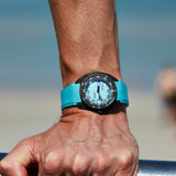 A close-up of a wrist wearing a DOXA SUB 300 Carbon Aquamarine Chronometer 42.50mm Watch 822.70.241.25 with a black bezel and a light blue dial, shown outdoors against a blurred beach background.