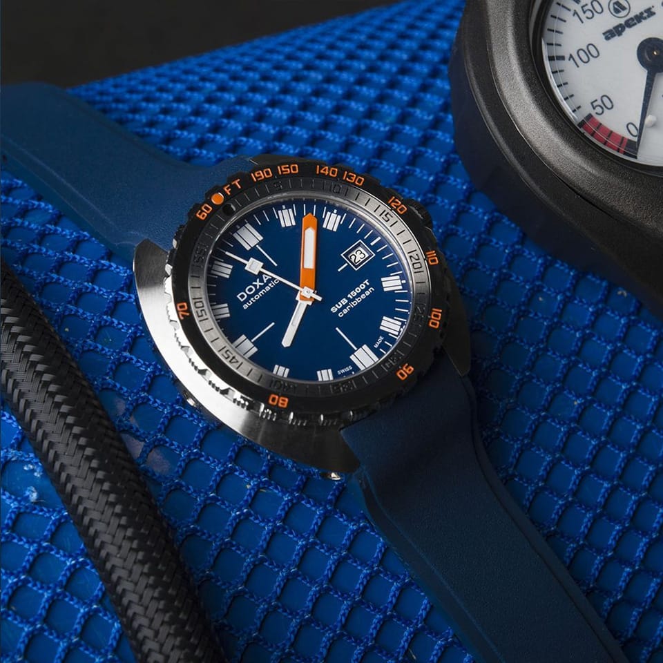 A close-up of a DOXA SUB 1500T Caribbean Automatic 45mm Watch 883.10.201.32watch with a blue dial, blue silicone strap, and black bezel, resting on a textured blue surface next to a carbon fiber strap.