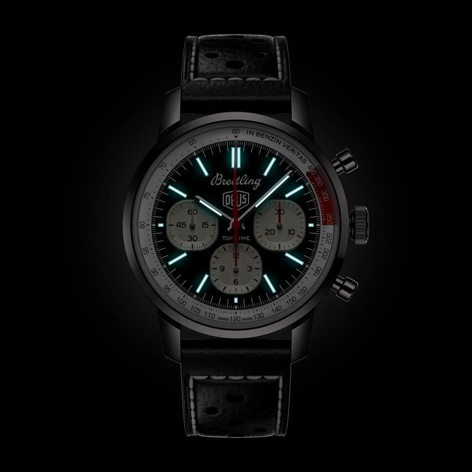 Breitling Top Time Deus B01 Chronograph 41mm Watch AB01765A1B1X1 shows the luminous properties of its dial markers and lightning-bolt-shaped chronograph hand in low light. The Deus emblem and squircle subdials remain legible against the illuminated black dial.