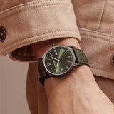 The Baume & Mercier Classima M0A10766 worn on a man's wrist in a lifestyle setting, showcasing the 42mm sandblasted stainless steel case with ADLC coating and green opaline dial catching the light, with the interchangeable green Holland & Sherry fabric strap sitting comfortably against the skin.