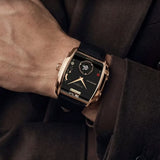 A close-up of a wrist wearing a luxury watch with a rectangular rose gold case, black dial, and black strap, styled with a dark brown suit jacket.
