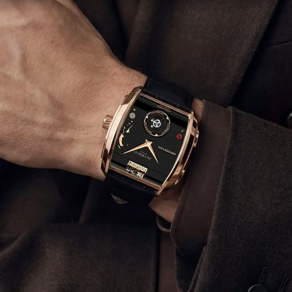 A close-up of a wrist wearing a luxury watch with a rectangular rose gold case, black dial, and black strap, styled with a dark brown suit jacket.