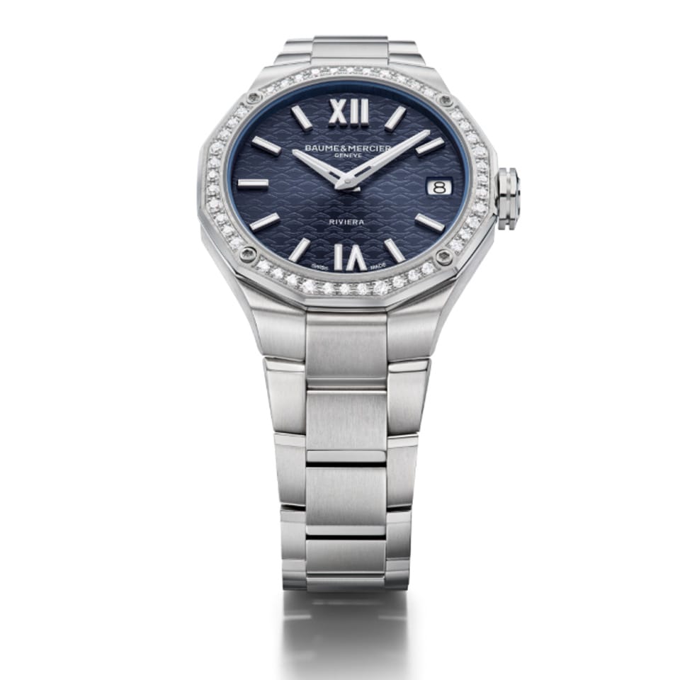 Overhead view of the Baume & Mercier Riviera M0A10765 ladies' watch, displaying the full watch face-up — the night blue dial with tone-on-tone wave transfers, riveted rhodium-plated Roman numerals and indexes, faceted rhodium-plated hands, and the sparkling 44 brilliant-cut diamond-set bezel catching the light, with the polished stainless steel bracelet laid out symmetrically on either side of the twelve-sided case.