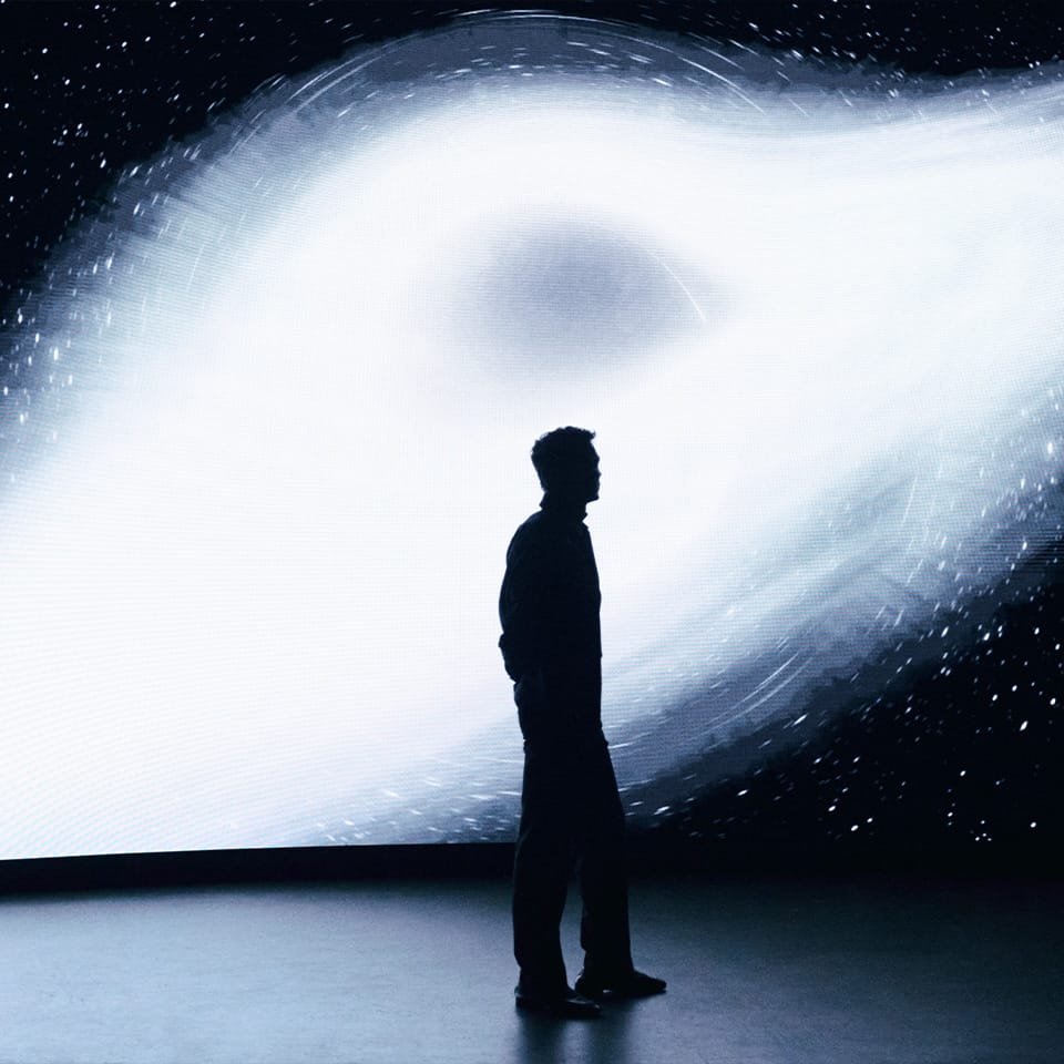 A person stands in silhouette facing a large glowing white and dark space display with swirling light effects, creating a dramatic high-contrast scene.