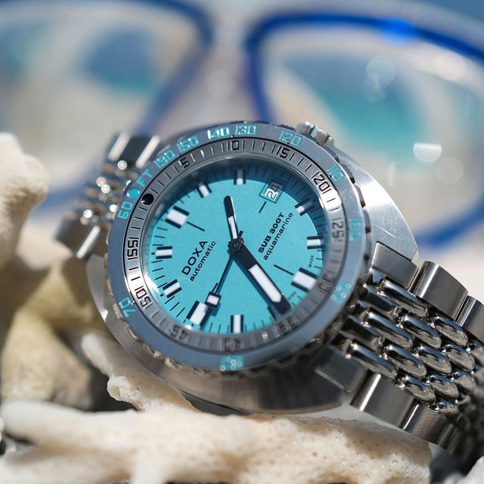 A DOXA SUB 300T Aquamarine Automatic 42.50mm Watch 840.10.241.10 with a bright turquoise dial and matching turquoise bezel markings, styled on white coral with a blurred blue-and-white diving mask in the background.