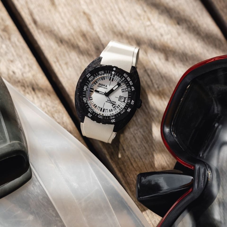 A DOXA SUB 300 CARBON Chronometer 42.50mm Watch 822.70.011.23 with a black bezel, white dial, and white rubber strap, styled on a wooden surface next to diving goggles.