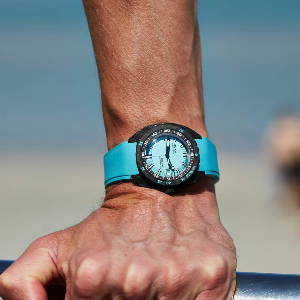 A close-up of a wrist wearing a DOXA SUB 300 Carbon Aquamarine Chronometer 42.50mm Watch 822.70.241.25 with a black bezel and a light blue dial, shown outdoors against a blurred beach background.