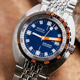 Close-up of a DOXA SUB 300T Caribbean Automatic 42.50mm Watch 840.10.201.10 wristwatch with a blue dial, orange accents, and a silver stainless steel link bracelet, set against a warm brown textured background.