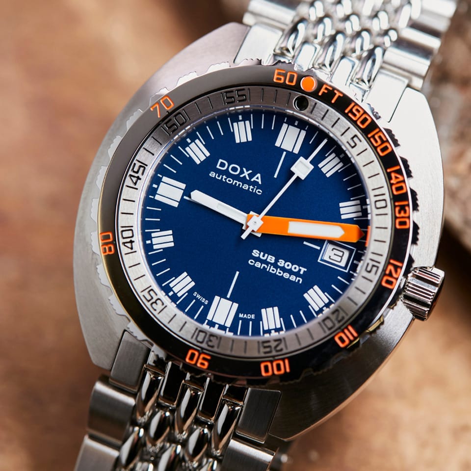 Close-up of a DOXA SUB 300T Caribbean Automatic 42.50mm Watch 840.10.201.10 wristwatch with a blue dial, orange accents, and a silver stainless steel link bracelet, set against a warm brown textured background.