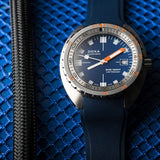 A DOXA SUB 1500T Caribbean Automatic 45mm Watch 883.10.201.32 watch with a blue dial, blue strap, and silver-toned case, displayed on a textured blue mesh surface next to a black braided strap.