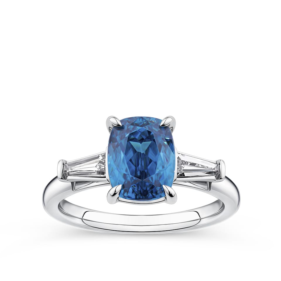 4.62 Carat Zircon and 0.36 Carat TW Diamond Trilogy Ring in 18ct White Gold — A 4.62 carat cushion-cut blue zircon sits at the centre of this trilogy design, flanked by two tapered baguette diamonds of G colour and VS clarity. The 18ct white gold setting provides a polished, cool contrast to the zircon's vivid blue hue.