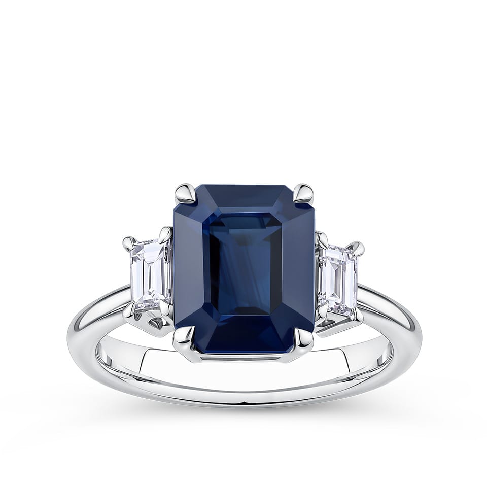 3.68 Carat Sapphire and 0.42 Carat TW Diamond Trilogy Ring in 18ct White Gold — A 3.68 carat emerald-cut blue sapphire sits at the centre of this claw-set trilogy design, flanked by two natural emerald-cut diamonds of G colour and VS clarity. The 18ct white gold setting provides a polished, cool contrast to the sapphire's deep blue hue.